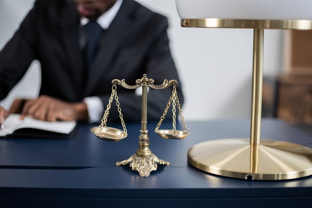 a-brass-balance-scale-sits-on-a-dark-blue-desk-next-to-a-gold-lamp-with-a-person-in-a-suit-writing-in-a-notebook-in-the-background-suggesting-a-legal-or-professional-setting